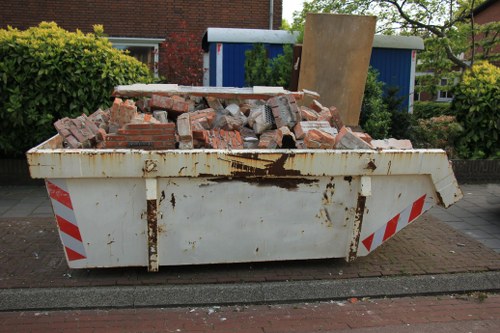 A large, white metal skip positioned on a paved sidewalk in a residential area, filled with a pile of reddish-brown bricks and rubble of varying sizes, some with rough, uneven surfaces and visible mortar residues. The skip shows signs of rust and weathering, with a rusty patch near the top edge. Behind the skip, there is a low hedge with green foliage, and further back, a blue and white utility shed or small building is partially visible. To the right, there's a brick building partially obscured by trees and shrubbery. The setting appears to be an urban or suburban street, indicative of rubbish removal or clearance services in Peckham, with natural daylight illuminating the scene and a peaceful, tidy environment typical of local waste management activity.