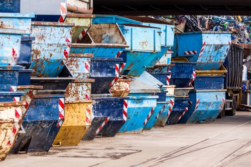 A row of multiple large industrial metal skips filled with assorted waste materials, positioned on a concrete pavement area, with signs of rust and wear on their surfaces. The skips are primarily painted in shades of blue, with some featuring white and red striped hazard markings along their edges. The waste inside appears to include scrap metal, packaging, and miscellaneous debris, seen spilling slightly over the sides. The environment suggests an outdoor waste management or rubbish disposal site, with a dark truck partially visible in the background to the far right, indicating that the skips are used for rubbish collection and removal services typically associated with local waste management in Peckham or nearby South London areas. The lighting is natural, highlighting the textures and weathered finishes of the skips, consistent with a professional rubbish removal setup operated by Peckham House Clearance or similar local services.