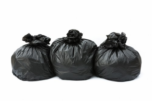 Three black plastic rubbish bags, filled and tightly knotted at the top, are lined up in a row against a plain white background. The bags appear to contain household waste and are made of thick, glossy plastic material with a slightly crinkled texture. They are positioned on a flat surface, possibly on a pavement or driveway, with minimal shadowing visible beneath them. The image depicts a typical scene of waste awaiting collection by a rubbish removal service in Peckham or surrounding areas, demonstrating the type of waste removal tasks that Peckham House Clearance services handle in residential or commercial settings. The bags are of similar size and shape, slightly leaning to the sides, emphasizing the bulkiness of the contents. This straightforward presentation highlights the common form of rubbish disposal before the waste is transported for disposal or recycling, fitting within the context of domestic or commercial clearance work in South London.