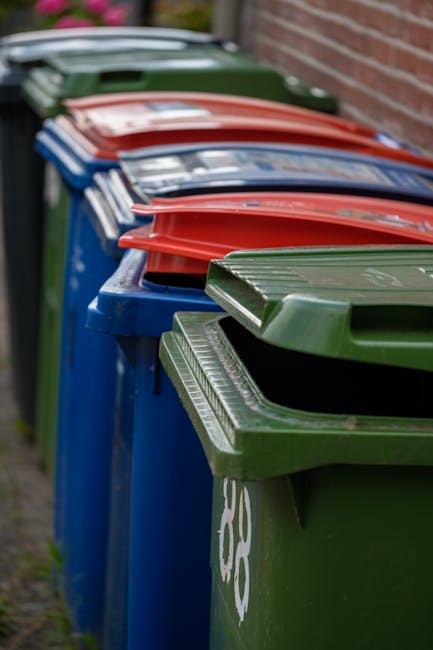 A street scene features a red rubbish collection truck with the rear hopper open, positioned on the roadside. A worker dressed in a high-visibility yellow and orange vest is standing behind the truck,