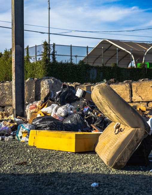 A pile of mixed waste materials accumulated on a gravel surface outdoors, including several black plastic rubbish bags, some partially torn open revealing contents like paper and plastic packaging, an