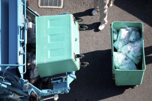 An overhead view of a waste collection scene in an outdoor area featuring two large green plastic bins, one of which is on a metal wheeled trolley, and the other filled with irregularly shaped plastic bags containing rubbish. The trolley bin is positioned next to a blue waste collection vehicle, partially visible in the left side of the image, with metallic components and hydraulic parts. A person, wearing light-colored trousers and white shoes, stands nearby on a dark asphalt surface, which has a rough texture. The environment suggests a typical street or alleyway in Peckham, with minimal surrounding context visible. The scene illustrates a rubbish removal process, accurately representing the type of waste and equipment used for house clearance services by Peckham House Clearance.
